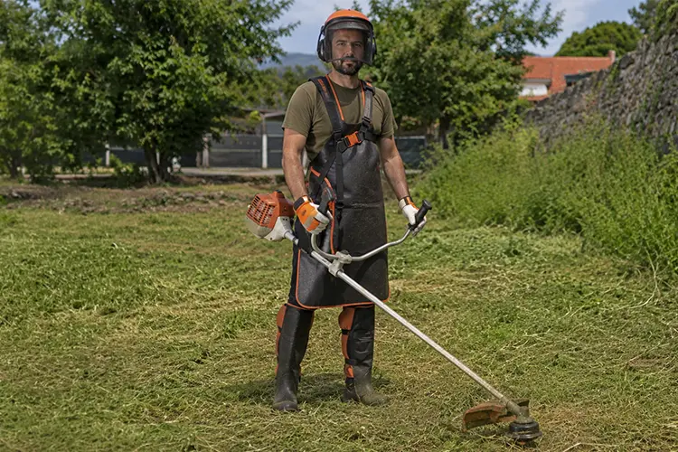 Exemplo de homem a usar o equipamento de segurança para roçadora: avental, viseira, proteção de ouvidos, luvas e caneleiras.